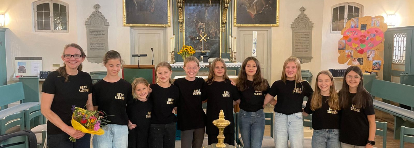 Einige Kinder des CHores stehen um einen Taufstein, sie tragen einheitliche Chorshirts